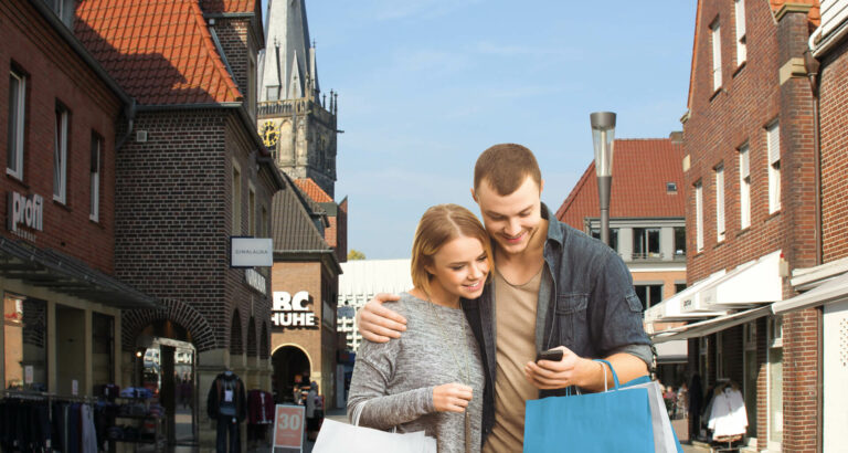 Junges Paar schaut lächelnd auf ein Smartphone, umgeben von Einkaufsstraßen mit Backsteingebäuden