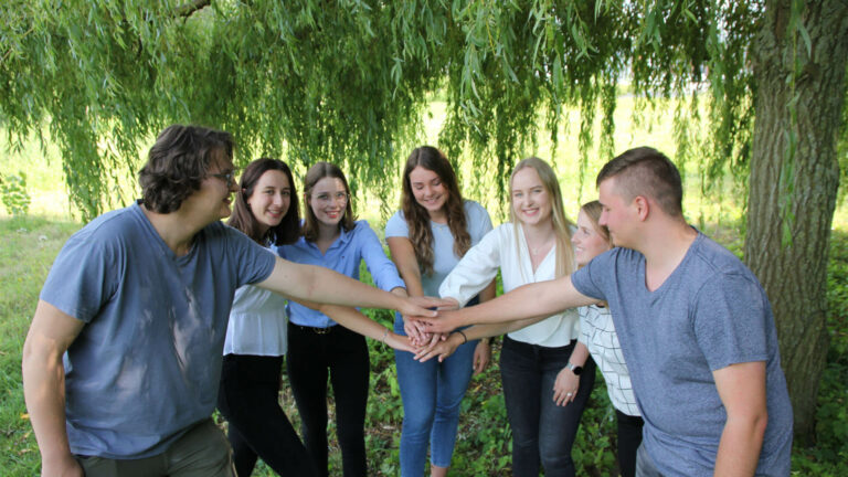 Sieben Personen stehen im Halbkreis und reichen jeweils eine Hand in der Mitte zusammen