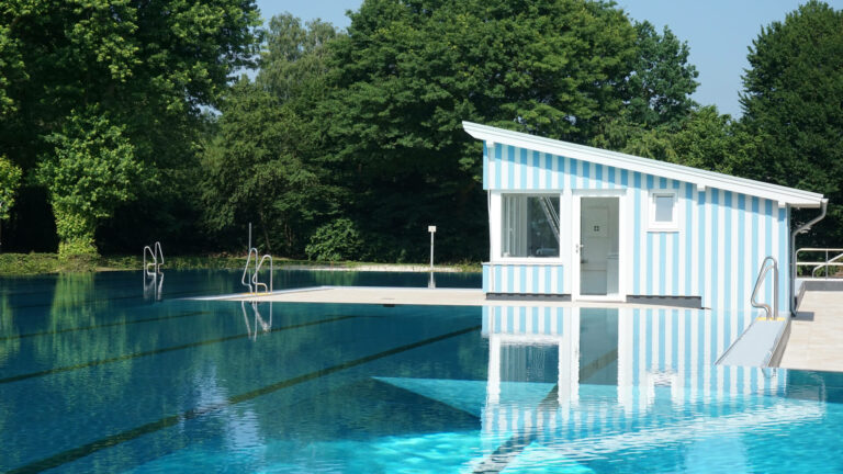 Freiluft-Schwimmbecken mit klarem Wasser, blaues gestreiftem Gebäude umgeben von grünen Bäumen