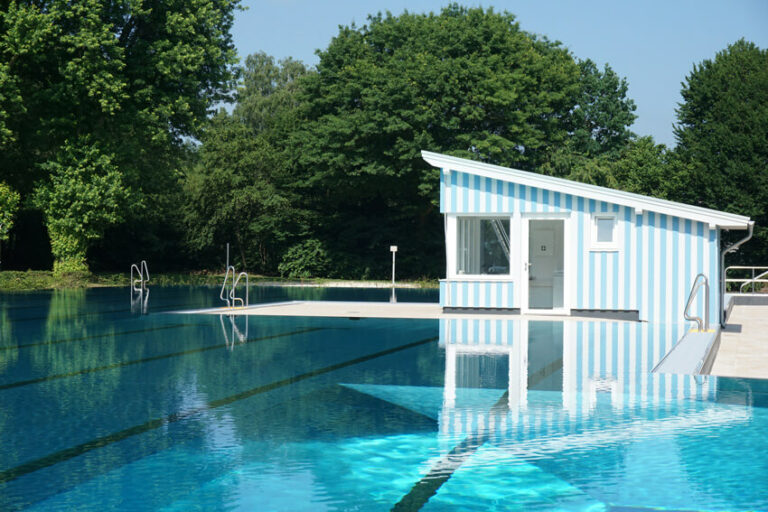 Freiluft-Schwimmbecken mit klarem Wasser, blaues gestreiftem Gebäude umgeben von grünen Bäumen