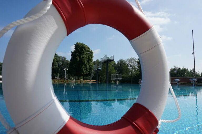 Rotes und weißes Rettungsring vor einem Schwimmbecken mit Sprungtürmen und Bäumen im Hintergrund