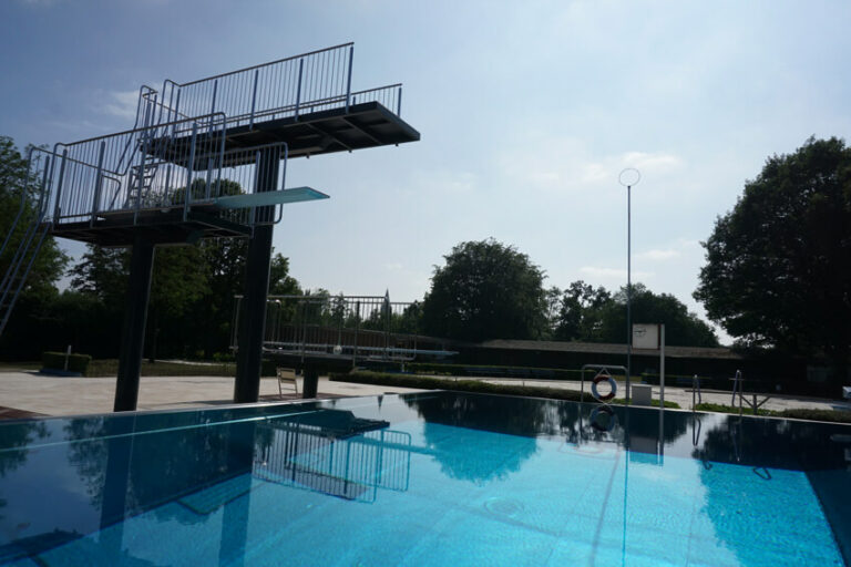 Leeres Schwimmbecken mit hohem Sprungbrett und Bäumen im Hintergrund unter bewölktem Himmel
