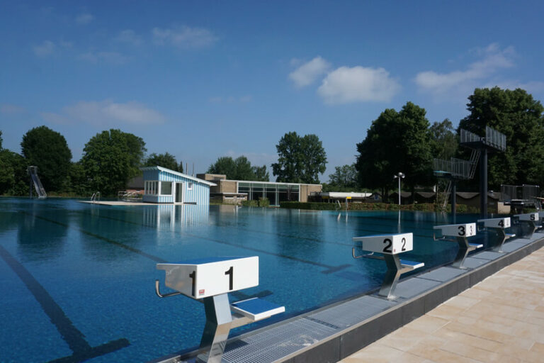 Leeres Freibad mit Startblöcken und Sprungtürmen bei klarem Himmel und umgeben von Bäumen
