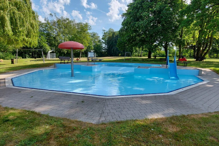 Kinderplanschbecken mit rotem Pilz-Wasserspender und blauer Rutsche mit Bäumen und Spielgeräten