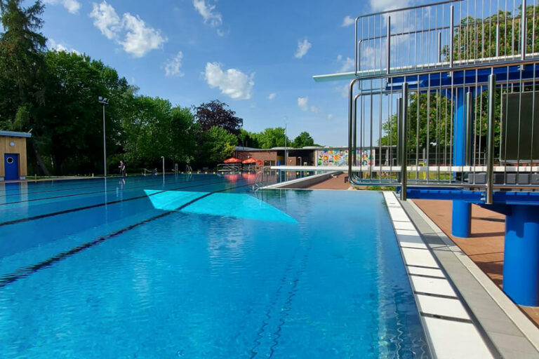 Freibad mit klarem Wasser, Sprungturm rechts und Bäumen im Hintergrund unter blauem Himmel