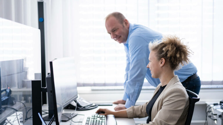 Kollegen im Büro, eine Frau sitzt am Schreibtisch vor dem Computer, ein Mann steht daneben