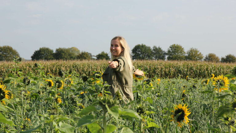 Frau mit heller Jacke steht lächelnd in einem Sonnenblumenfeld und zeigt nach vorne