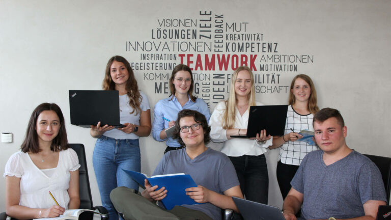 Sieben junge Erwachsene, einige mit Laptops und Notizbüchern, vor einer Wand mit Schrift