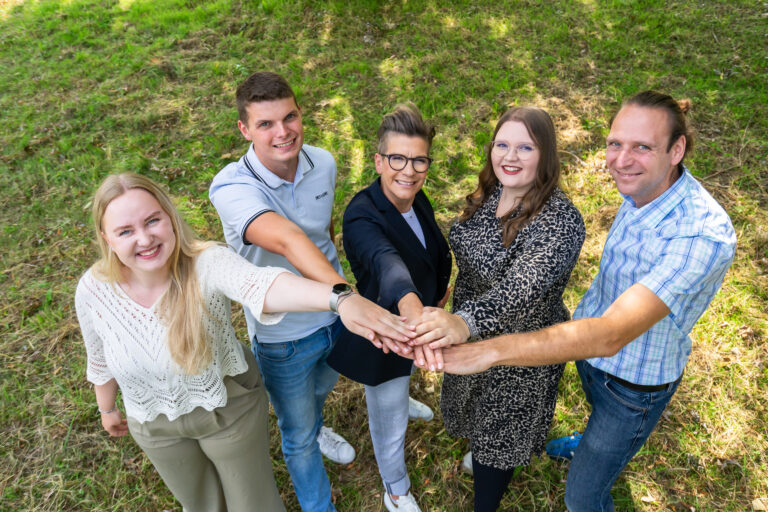 Fünf Personen führen alle eine Hand zusammen und schauen in die Kamera