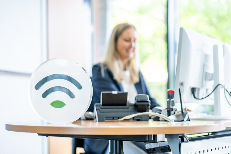Frau arbeitet am Computer in einem Büro, im Vordergrund ein Tisch mit einem WLAN-Symbol