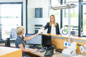 Empfangsmitarbeiterin übergibt einer lächelnden Besucherin an der Rezeption ein Dokument