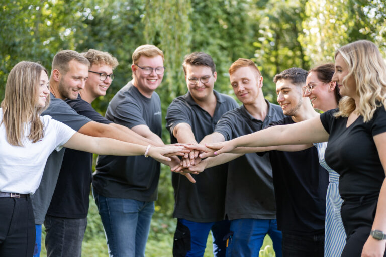Acht Personen stehen im Halbkreis und reichen jeweils eine Hand in der Mitte zusammen