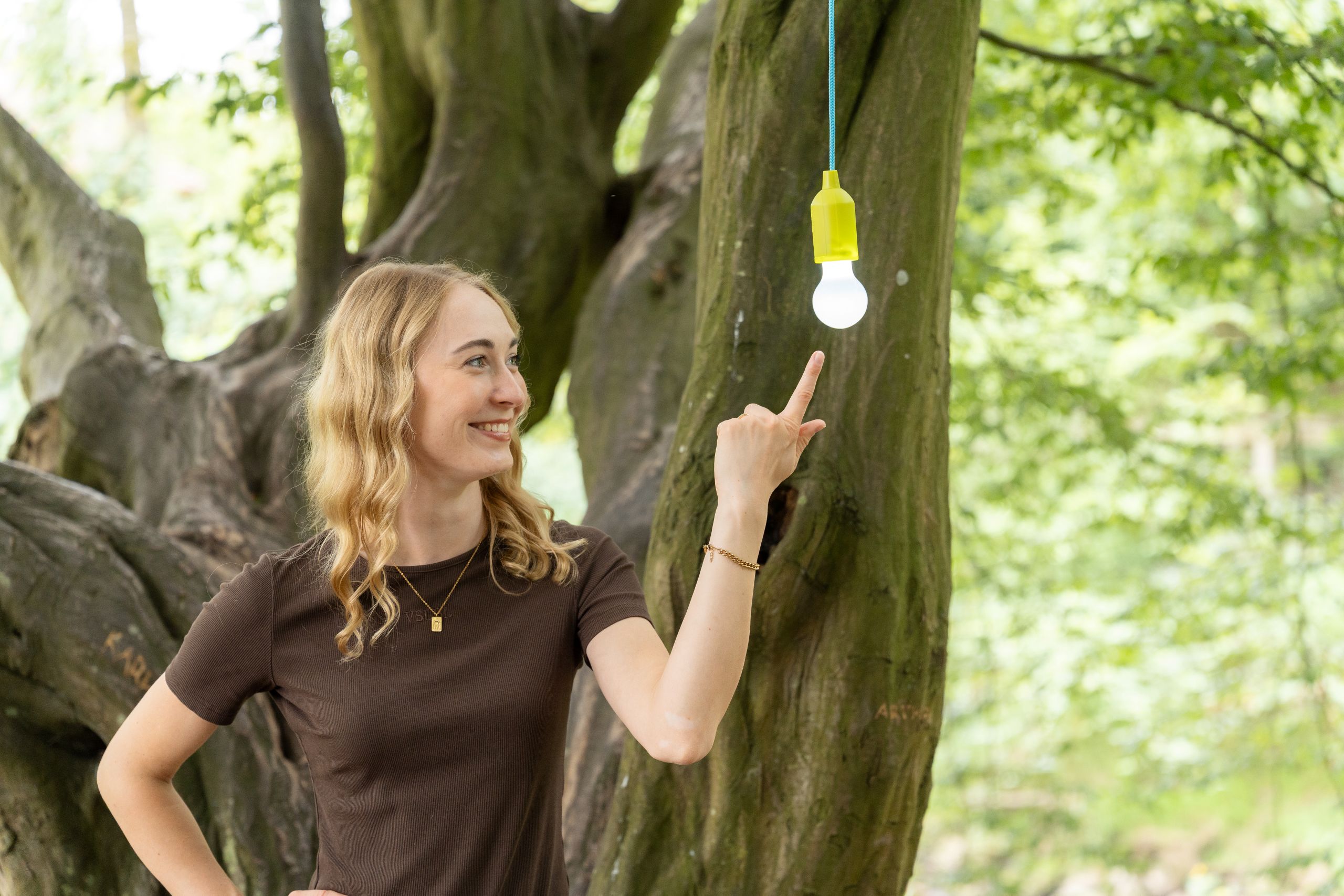 Eine Frau steht vor einem Baum und zeigt auf eine leuchtende Glühbirne die vom Baum hängt.